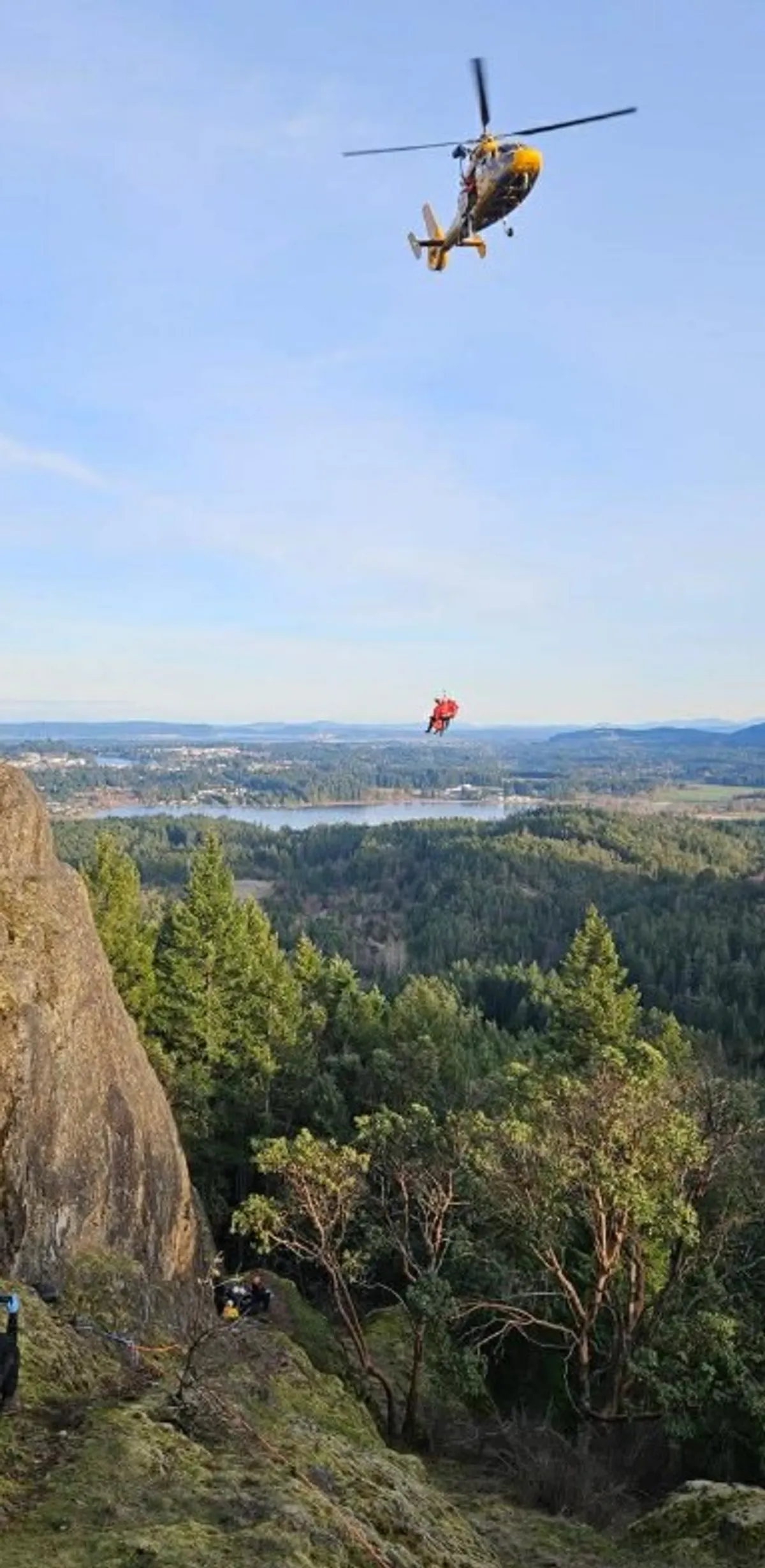 SAR crews accompany an injured rock climber as he's hoisted into a North Shore Rescue helicopter.
